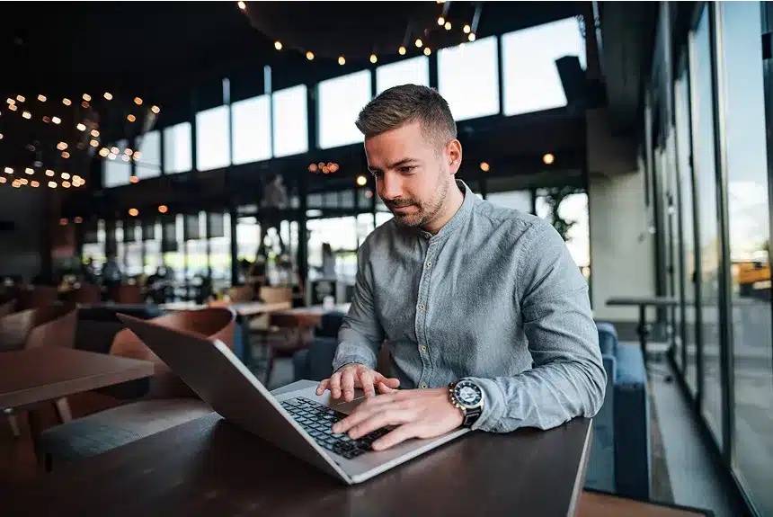 A man focused on his laptop while seated at a table in a bustling restaurant.