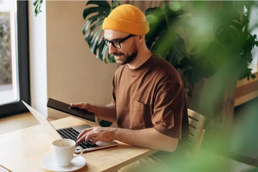 A man wearing a beanie sits at a table, focused on his laptop.