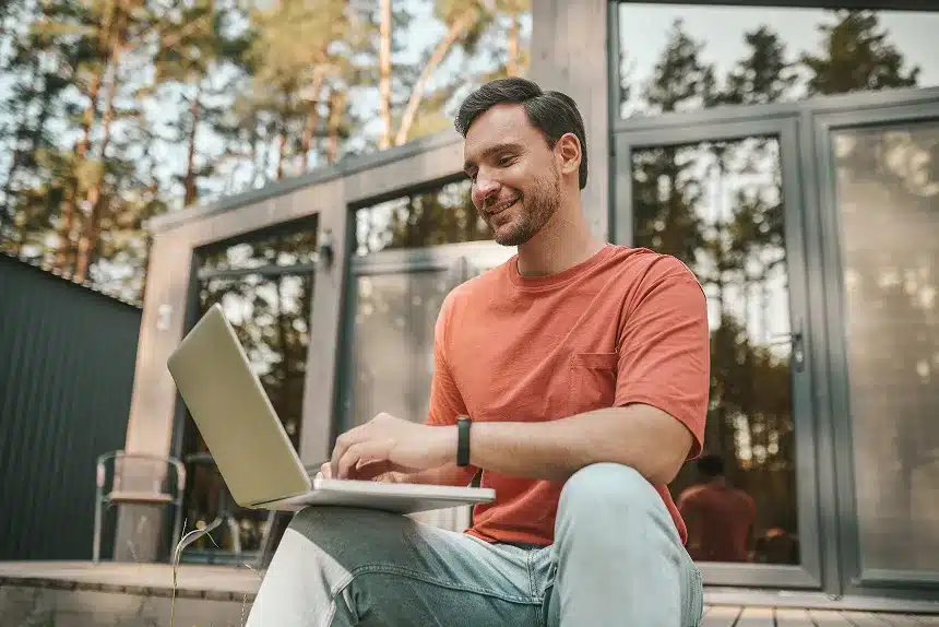 A man sitting on a porch, working on a laptop, surrounded by a peaceful outdoor setting.