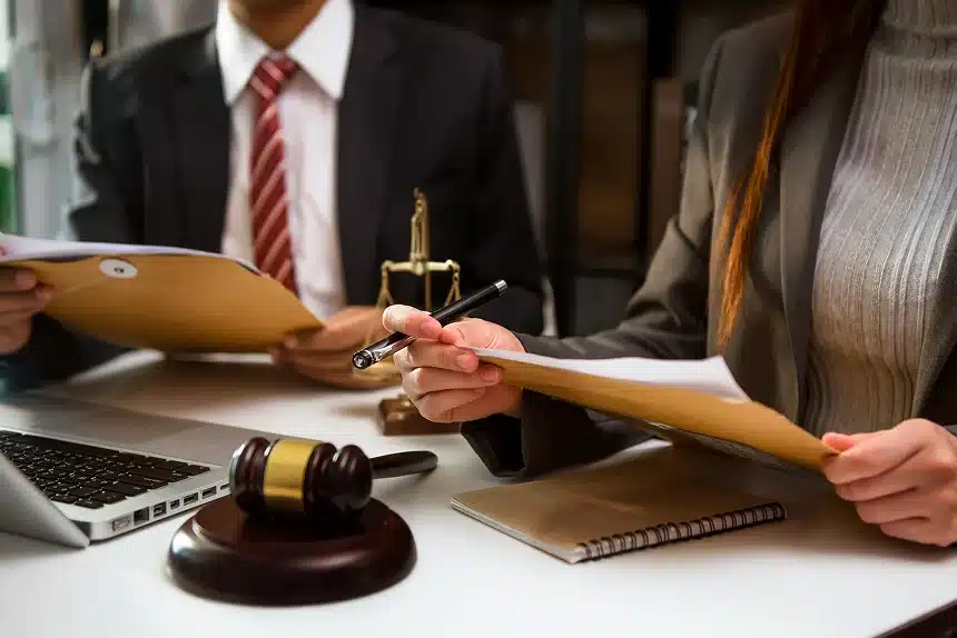 Two people in business attire review documents. A gavel, notebook, and laptop are on the desk, suggesting a legal or corporate setting. Serious tone.