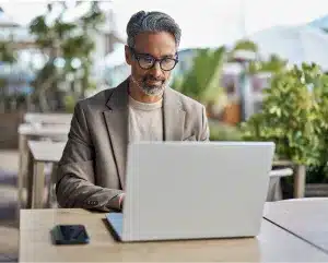 A man wearing glasses is seated at a table, focused on his laptop.