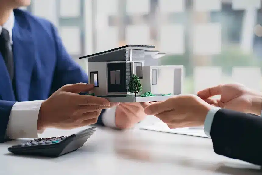 Two individuals are holding a small model of a house, smiling and standing together in a bright, well-lit environment.
