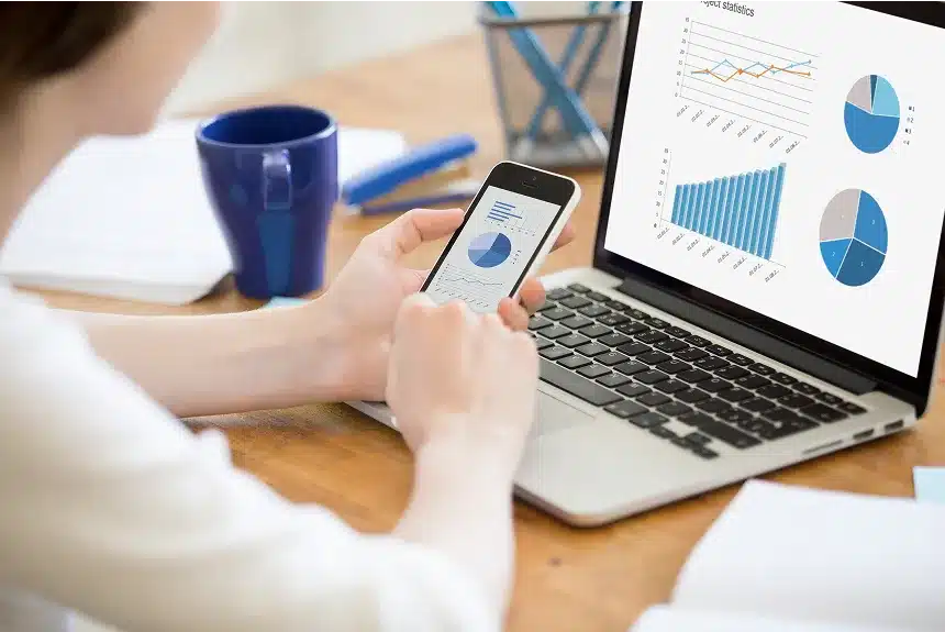 A woman examines a graph on her laptop while holding her phone in her other hand.