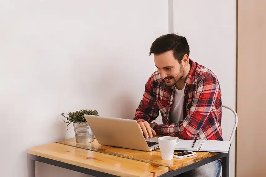 A man sitting at a table, focused on his laptop, with a neutral background.
