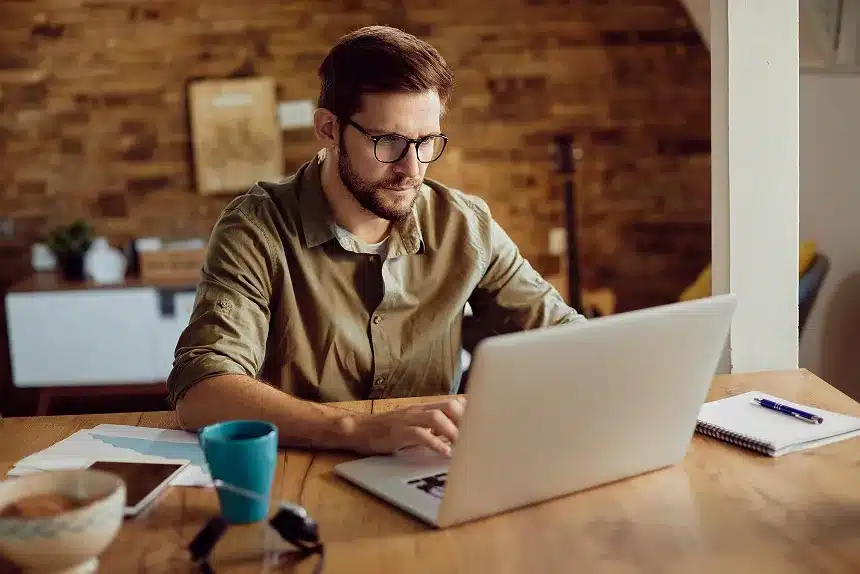 A man wearing glasses sits at a table, focused on his laptop while working or studying.