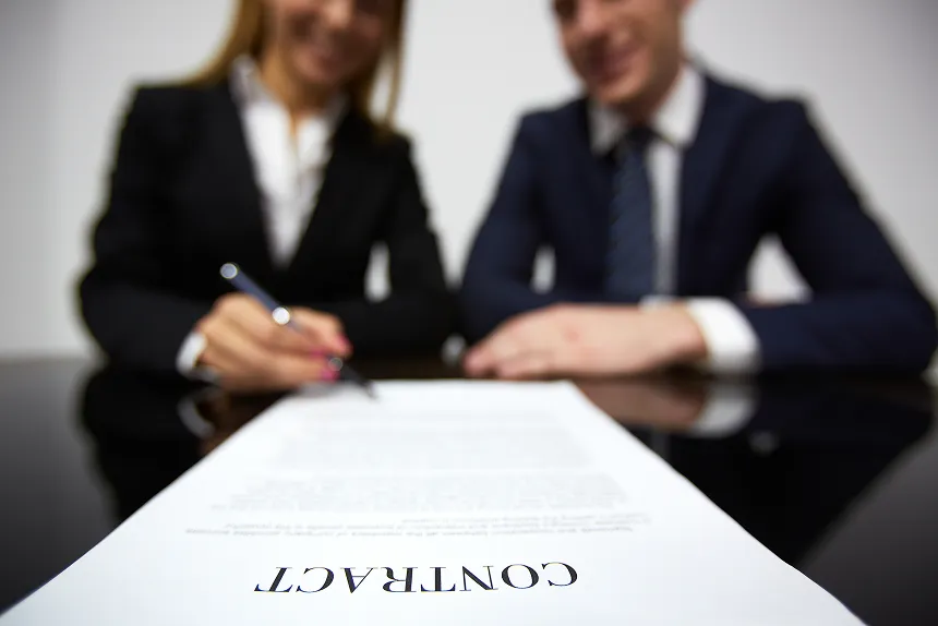 A business couple signing a contract at a table, with documents and pens in front of them.