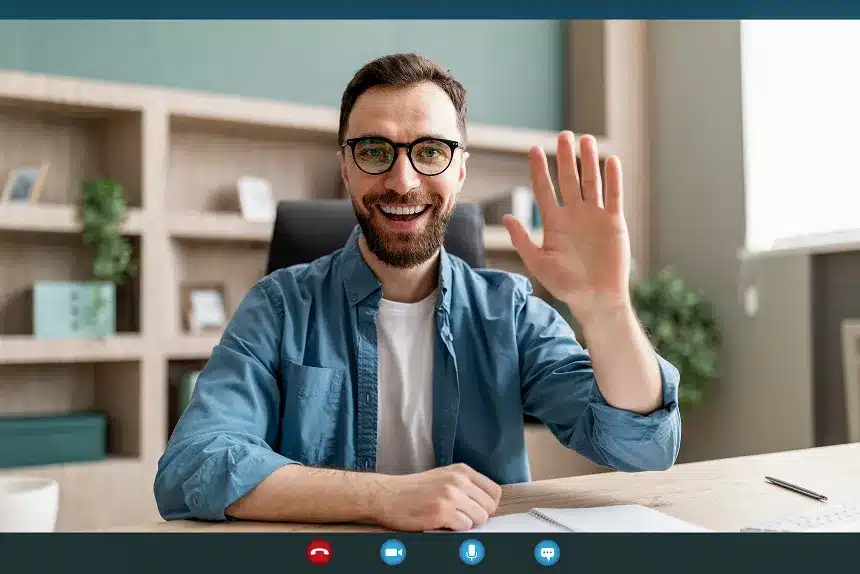 A man with glasses is smiling and waving at the camera, creating a friendly and inviting atmosphere.