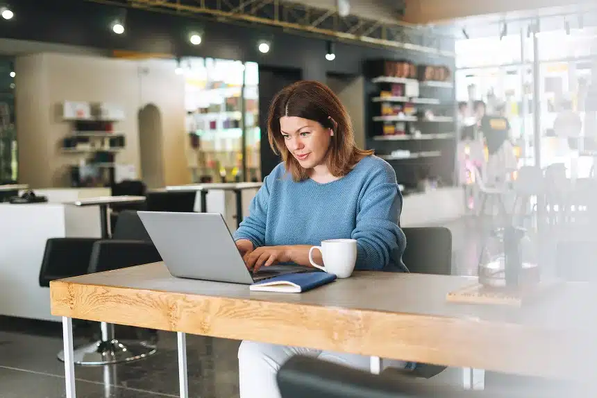 A woman seated at a table, focused on her laptop, with a calm expression in a well-lit room.
