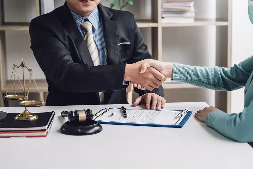 Two people shaking hands across a desk in an office setting. A lawyer in a suit sits beside a gavel and scales of justice, with documents on the table.