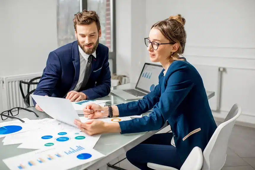 A man and a woman in business attire discuss charts and graphs at a desk. The scene conveys a professional, collaborative atmosphere.