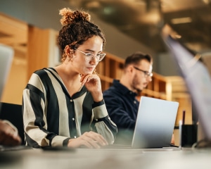 A woman wearing glasses is focused on her laptop, engaged in work at a well-lit desk.