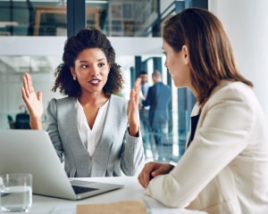 Two businesswomen engaged in a discussion at a table, exchanging ideas in a professional setting.