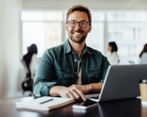 A smiling man wearing glasses sits at a desk, working on a laptop, exuding a sense of contentment and focus.