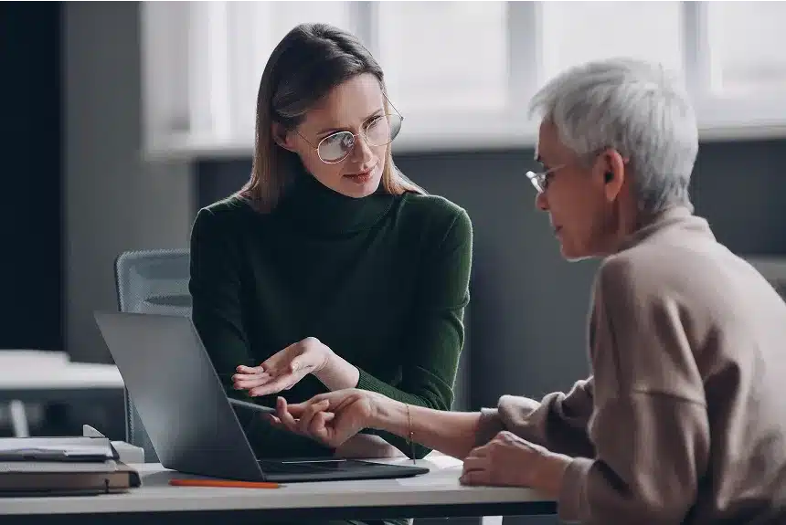 A woman and an older man are seated at a table, engaged in conversation while using a laptop.