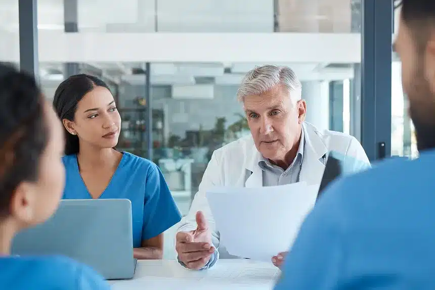 A doctor converses with a patient while two nurses attentively listen in a clinical setting.
