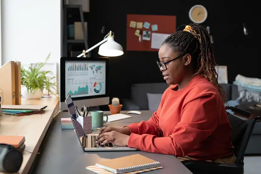 A woman in a red shirt sits at a desk, focused on her laptop while working.