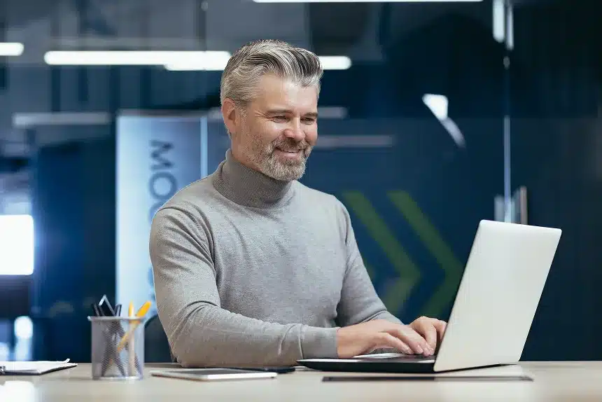 A man in a sweater sits at a desk, focused on his laptop, with a neutral background.