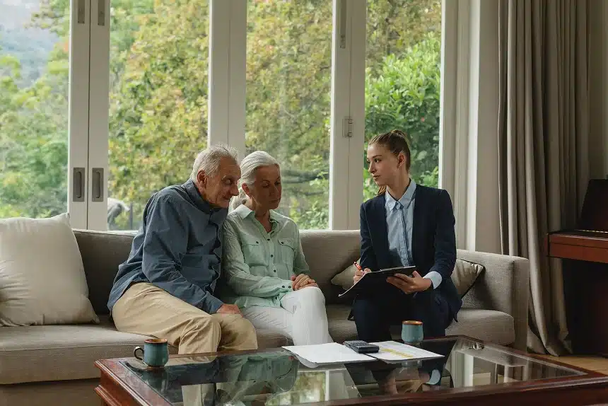 An elderly couple sits on a sofa in a bright room, attentively listening to a woman in a suit holding a clipboard. The atmosphere is calm and professional.