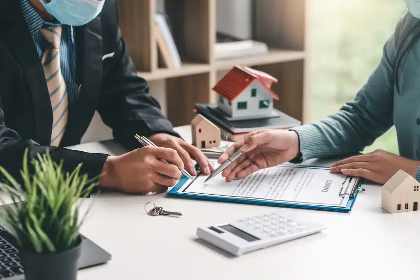 Two people in business attire and masks review a contract at a desk with a small house model. A pen, keys, and calculator are visible, suggesting a real estate transaction.
