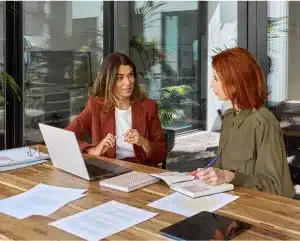 Two women engaged in conversation while working on a laptop at a table, showcasing collaboration and productivity.