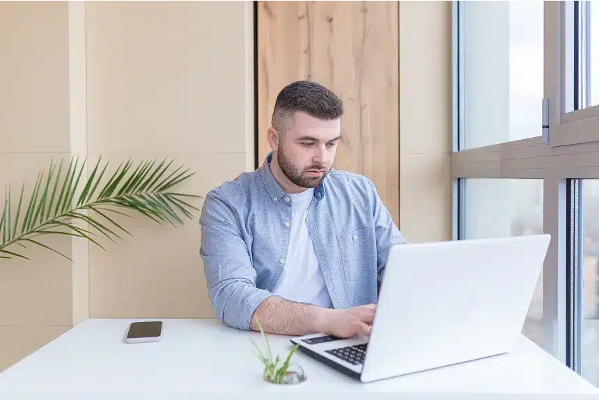 A man sitting at a desk, focused on his laptop, with a notepad and pen beside him.