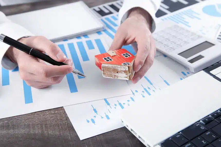A man's hand holds a house model on a desk with a laptop and calculator, symbolizing real estate or financial planning.