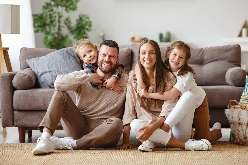 A family of four sitting on the floor, smiling and playing together in front of a cozy couch.