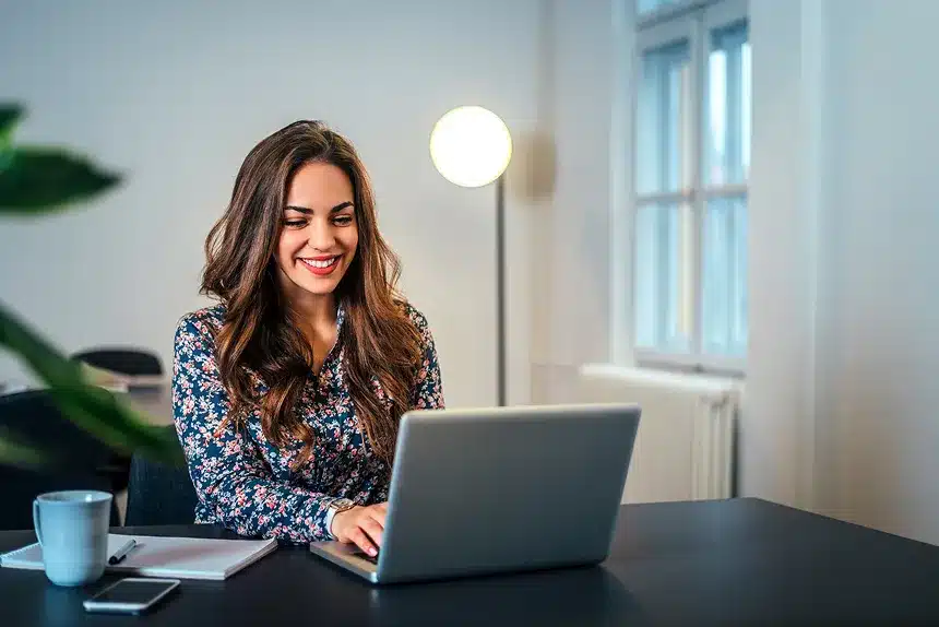 A woman smiles while working on her laptop, showcasing a positive and engaged expression.