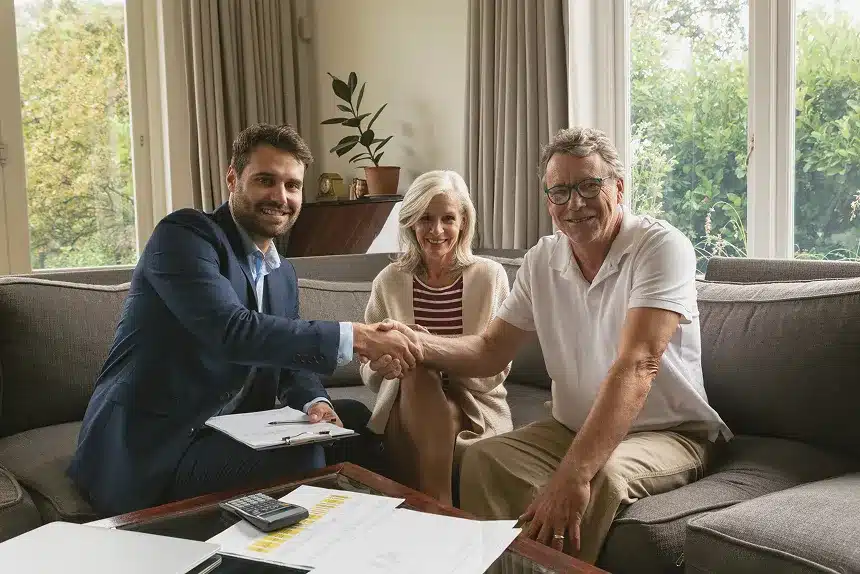 A man and woman shake hands while another couple sits on a couch, engaged in conversation.
