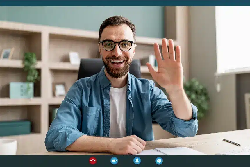 A man wearing glasses is smiling and waving at the camera.