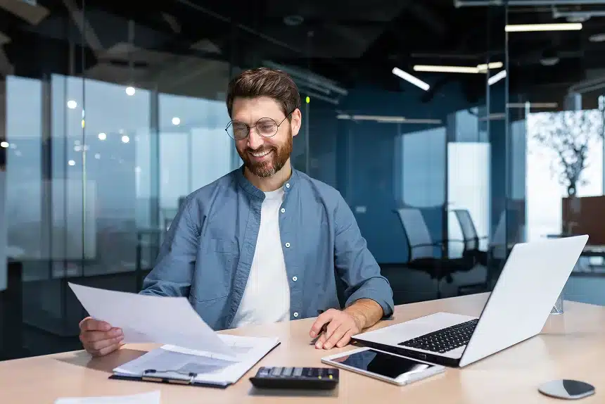 A bearded man sits at a desk, working on a laptop surrounded by scattered papers.