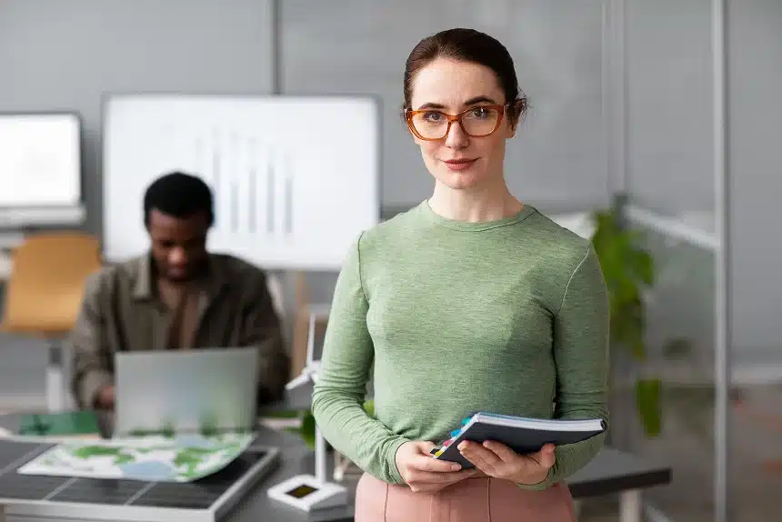 A woman wearing glasses stands in front of a desk with a laptop, appearing focused and engaged in her work.