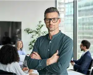 A man wearing glasses stands confidently in front of a diverse group of people engaged in conversation.