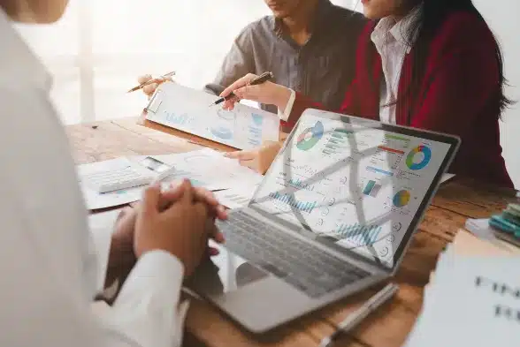 Three individuals seated at a table, engaged with a laptop and scattered papers, collaborating on a project.
