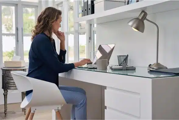 A woman seated at a desk, focused on her laptop, surrounded by a tidy workspace and soft lighting.