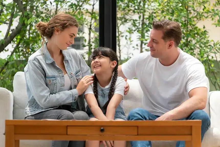 A man and woman sit on a couch, smiling at a child who is playing between them.