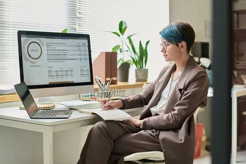 A woman sitting at a desk, working on a laptop with a desktop computer beside her.