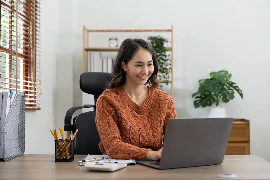 A woman sitting at her desk, focused on her laptop, with a notepad and pen beside her.