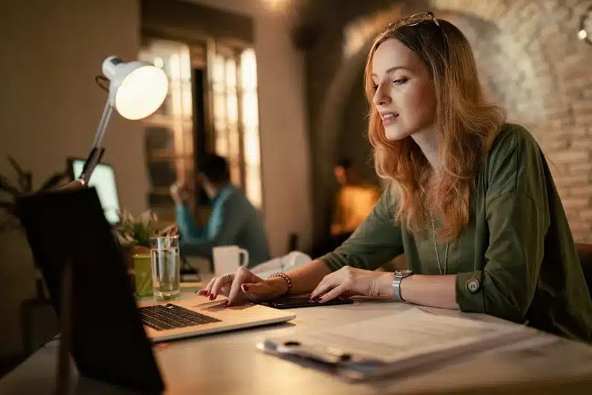 A woman is focused on her laptop while seated at a desk, surrounded by office supplies and a plant.