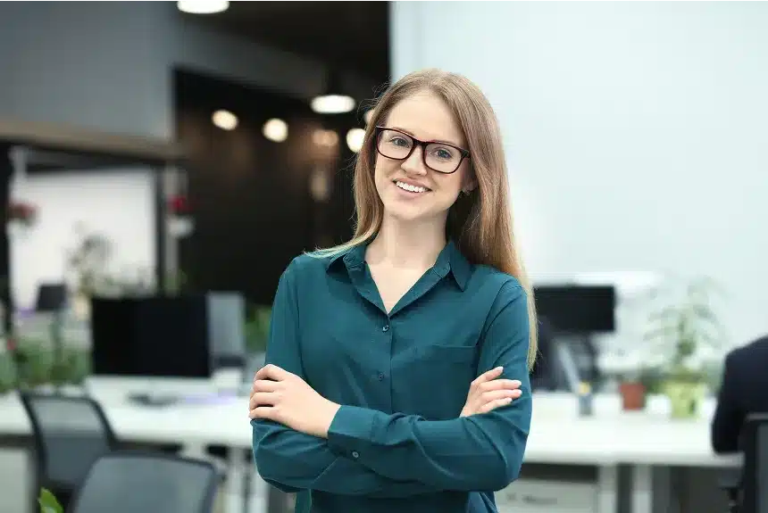 A woman wearing glasses stands in an office, surrounded by desks and office equipment, looking confidently at the camera.