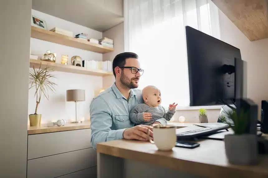 A man wearing glasses sits at a desk with a baby on his lap, both engaged in a playful interaction.