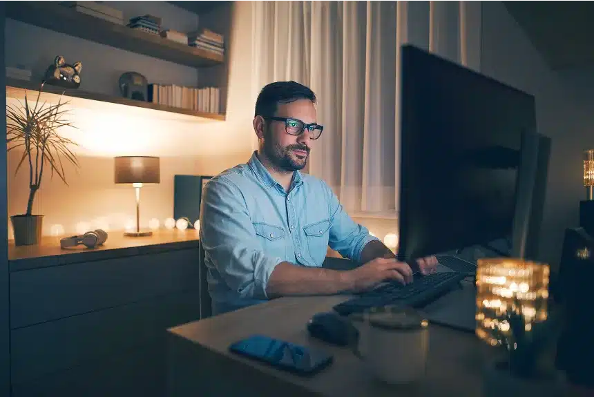 Man wearing glasses focused on working on a computer at home, surrounded by a cozy and organized workspace.