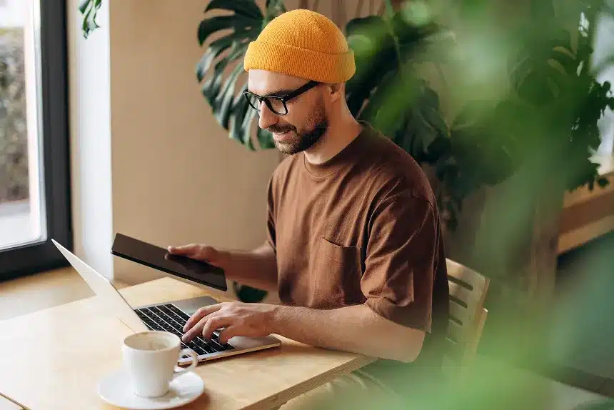 A man wearing a beanie sits at a table, focused on his laptop.