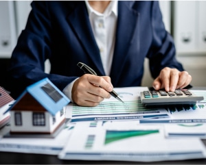 Businesswoman calculating finances with a calculator and a house model on her desk, focused on her work.