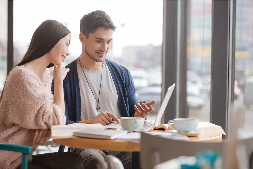 A man and woman sitting at a table, focused on a laptop, engaged in discussion or collaboration.