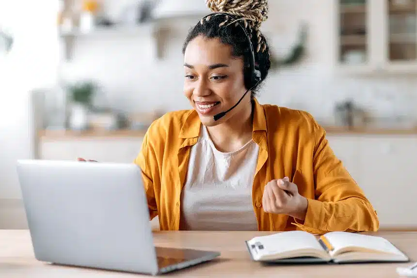 A woman with a headset sits at a table, focused on her laptop, engaged in a virtual meeting or work session.