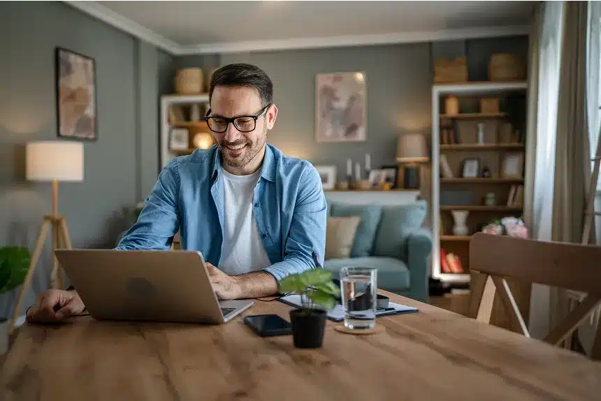 A man wearing glasses sits at a table, focused on his laptop while working or studying.