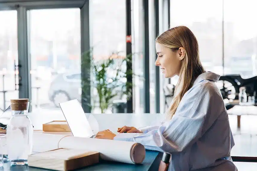 A woman sitting at a table, focused on her laptop, with a calm expression in a well-lit room.