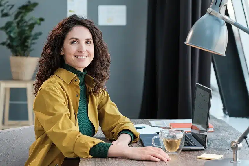 A woman sitting at a desk, focused on her laptop, with a notepad and pen beside her.