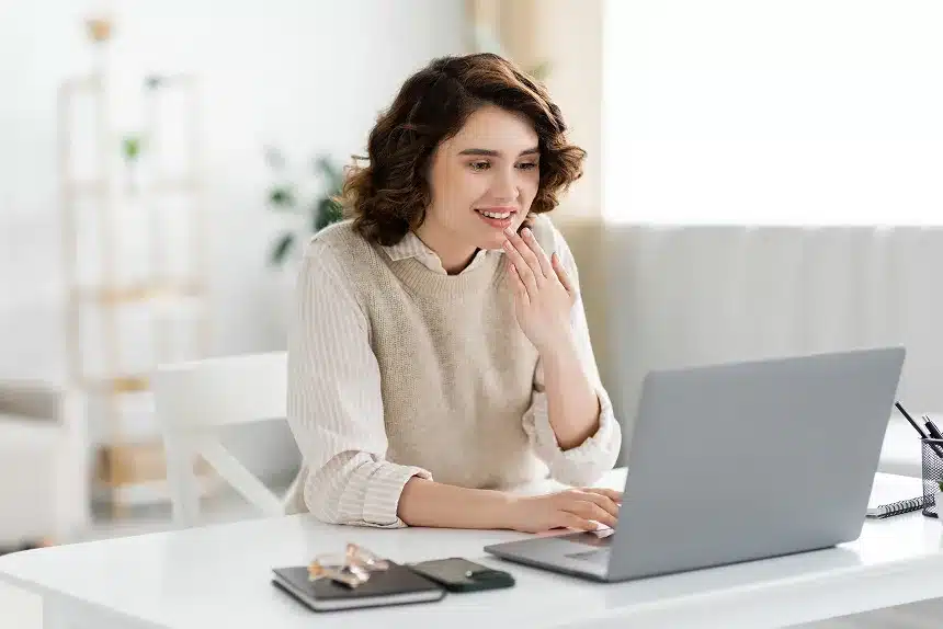 A woman sits at a desk, focused on her laptop, with a notepad and pen beside her.